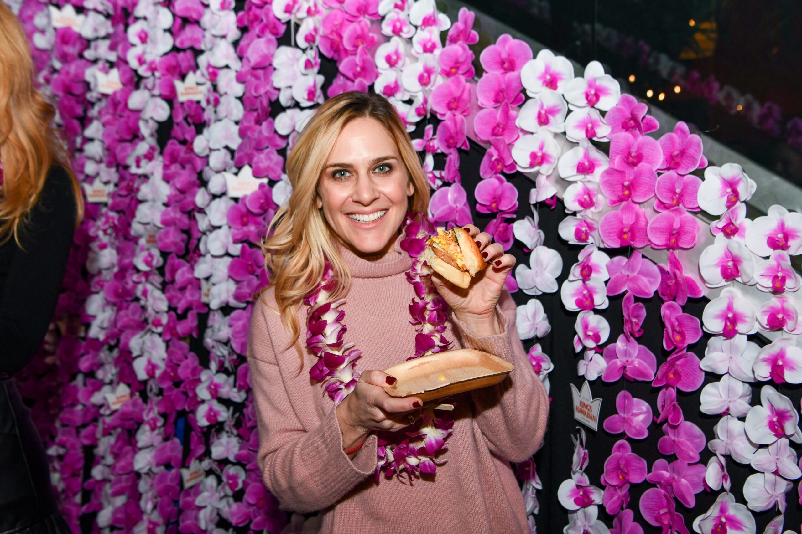 A guest posing in front of a wall of leis with a breakfast sandwich.
